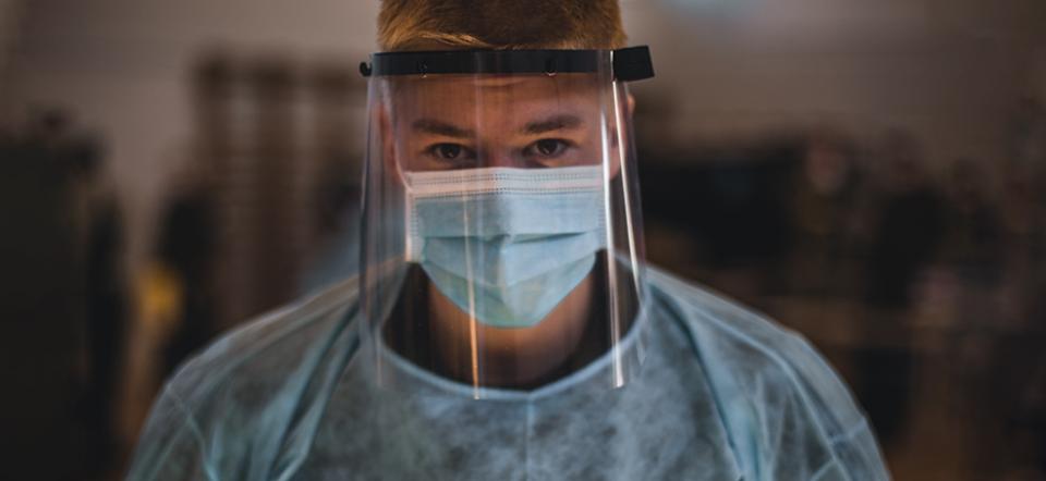 Man stands wearing medical equipment in the form of a mask, visor and suit.