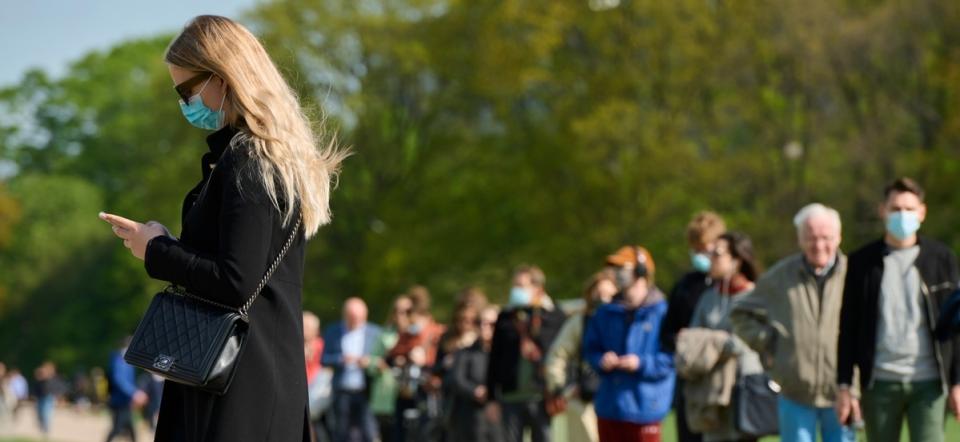 Folk står i kø med mundbind på. En kvinde i forgrunden tjekker sin mobil.