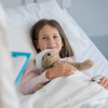 Pige ligger i en hospitalsseng med en bamse i hånden.
